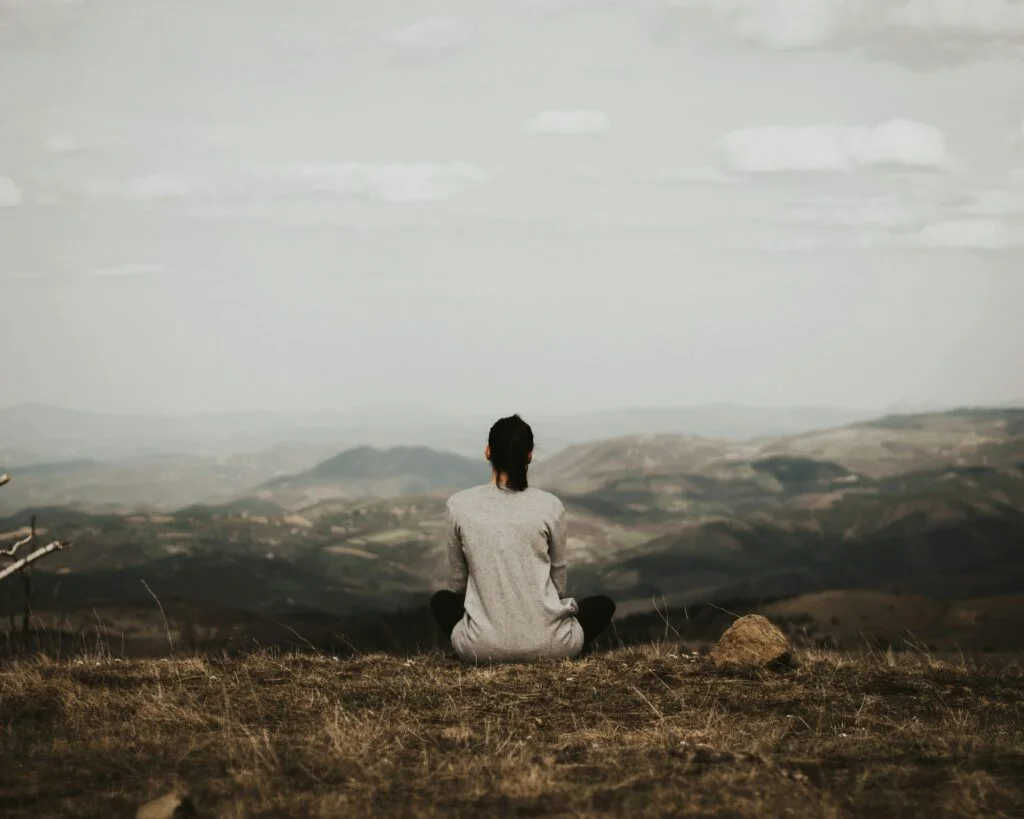 A woman sits cross legged on top of a hill overlooking fields, hills and a small village. The sky is gray and overcast.