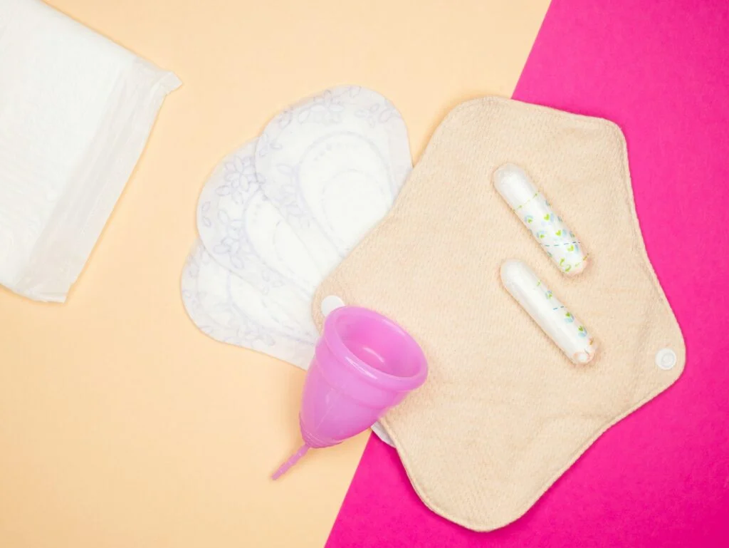 A brown reusable period bad, two tampons, one silicone menstrual cup, three panty liners and one overnight pad sit on a surface that's brown and pink.
