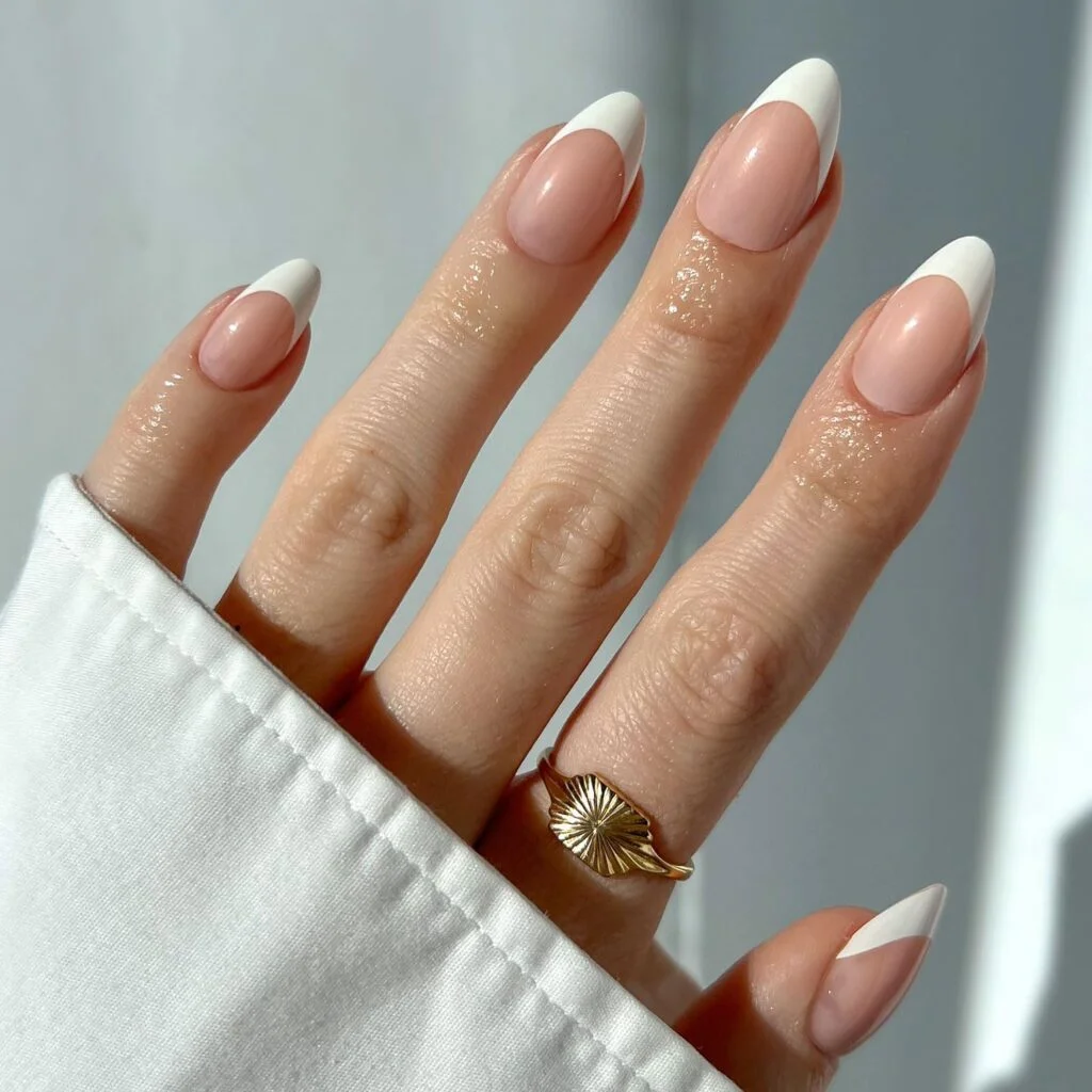 A woman wears a pair of French tip Glamnetic press-on nails with a gold ring and a white long sleeved shirt.