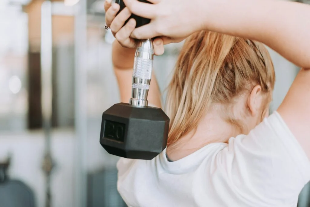 A blonde woman in a white short sleeved shirt lifts a dumbbell over her head, behind her neck.