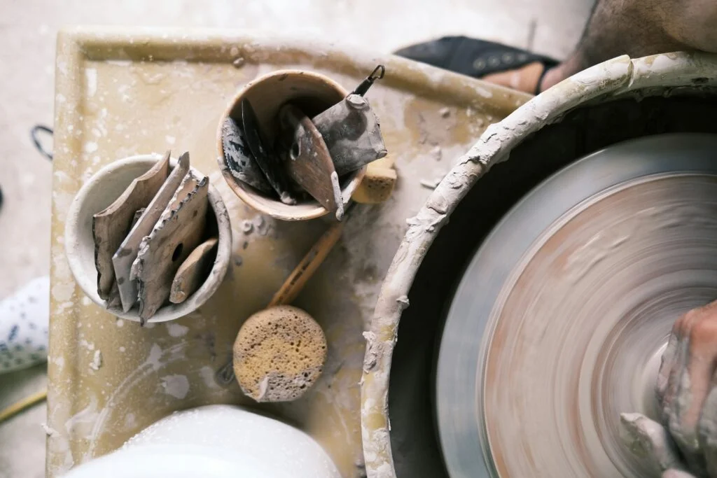 A potter's hand shapes clay on a spinning wheel. Nearby are a wet sponge and tools in cups on a dusty surface, conveying a sense of creativity and focus.