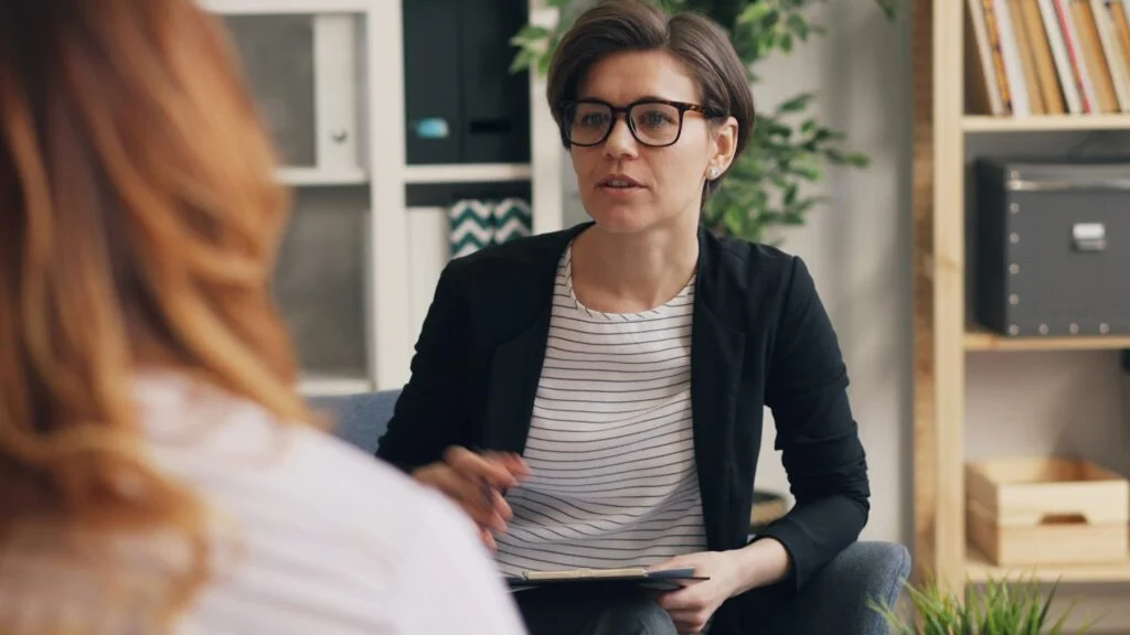 A female therapist with glasses and short hair, dressed in a black blazer and white and black striped shirt, attentively listens and takes notes during a serious discussion in an office setting.
