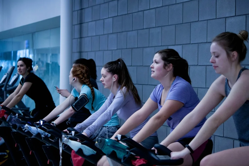 Six women in workout attire focus intently while cycling on stationary bikes in a gym. They wear earbuds, conveying determination and concentration.