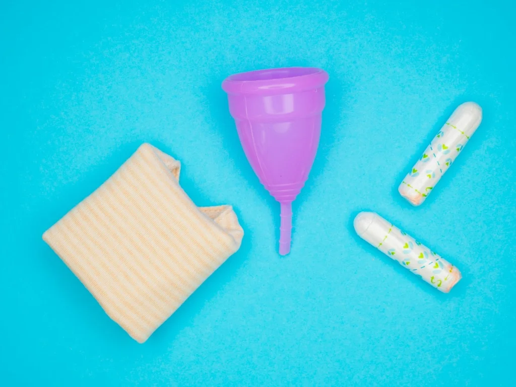 Menstrual products on a bright blue background: a folded beige pad, a purple menstrual cup, and two white tampons with patterned wrappers.