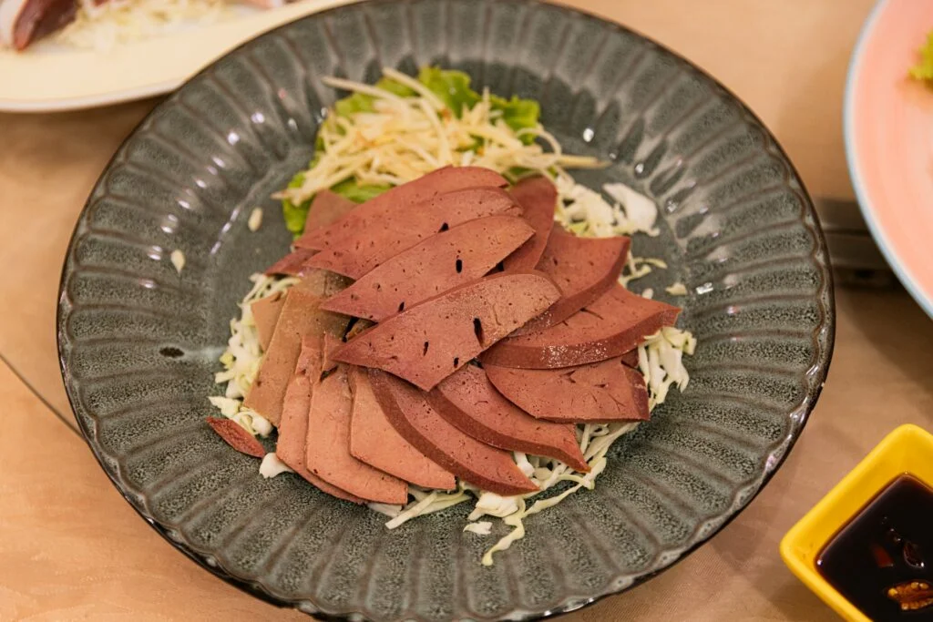 Sliced cooked liver on shredded cabbage and greens in a dark gray plate, accompanied by a small yellow dish of dipping sauce on the side.