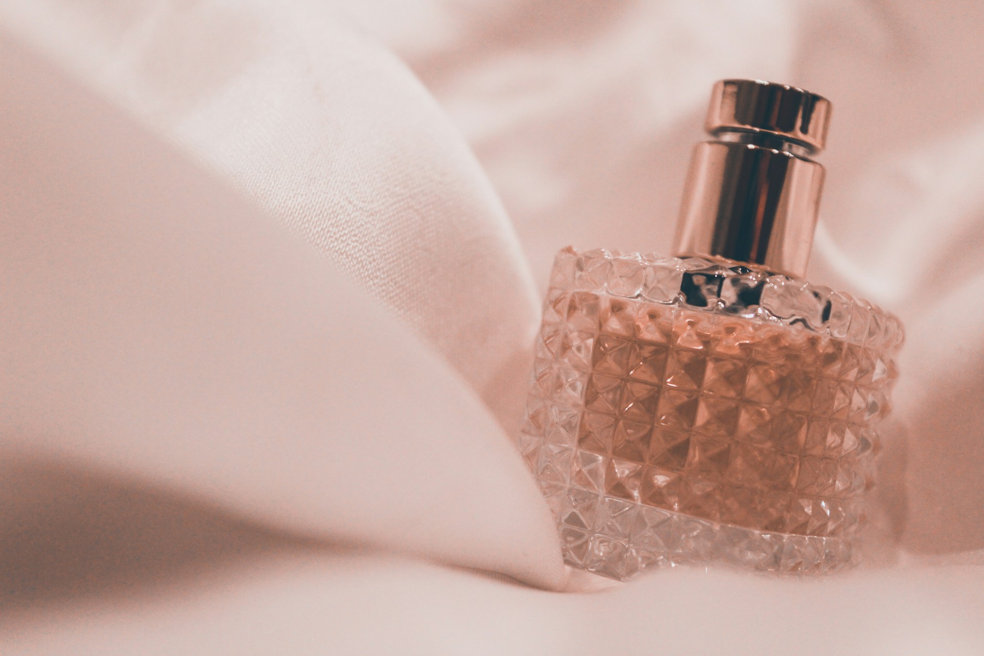 A crystal-patterned glass perfume bottle with a gold cap rests on top of soft, white luxurious fabric.