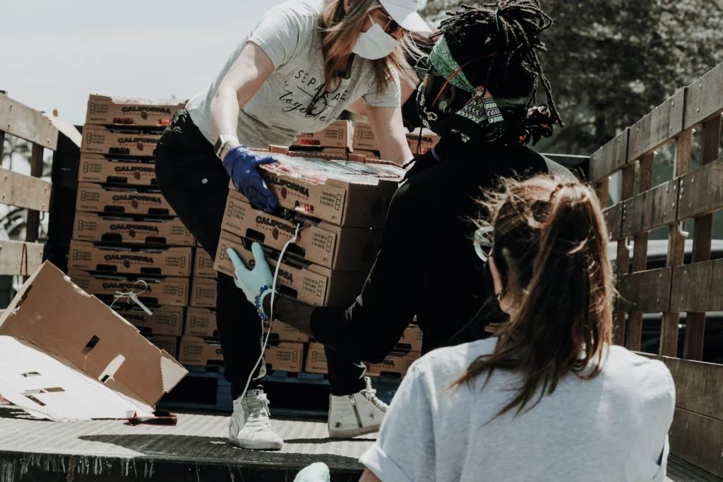 Three people, wearing masks and gloves, work together unloading boxes from a truck. The scene conveys teamwork and a community support effort.