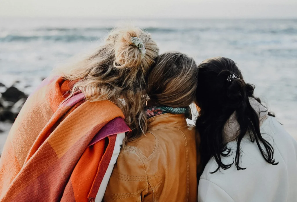 Three friends stand closely, facing the ocean with their backs to the camera. They wear warm jackets, conveying a sense of warmth and companionship by the sea.
