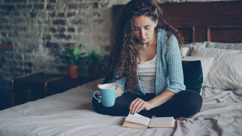 A woman with curly hair reads a book on a bed while holding a blue mug. The setting is cozy, with plants and a brick wall in the background.