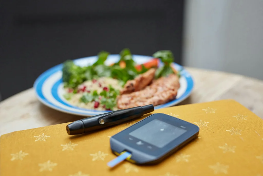 A glucose meter and lancet pen rest on a yellow surface in the foreground. In the background, a plate of vegetables and grilled meat is visible, suggesting a balanced meal.