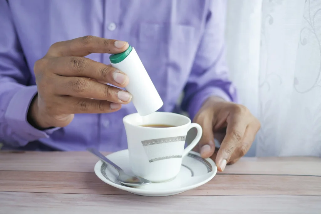A person in a purple shirt adds sweetener to a cup of tea on a saucer, with a metal spoon beside it, evoking a calm, morning routine.