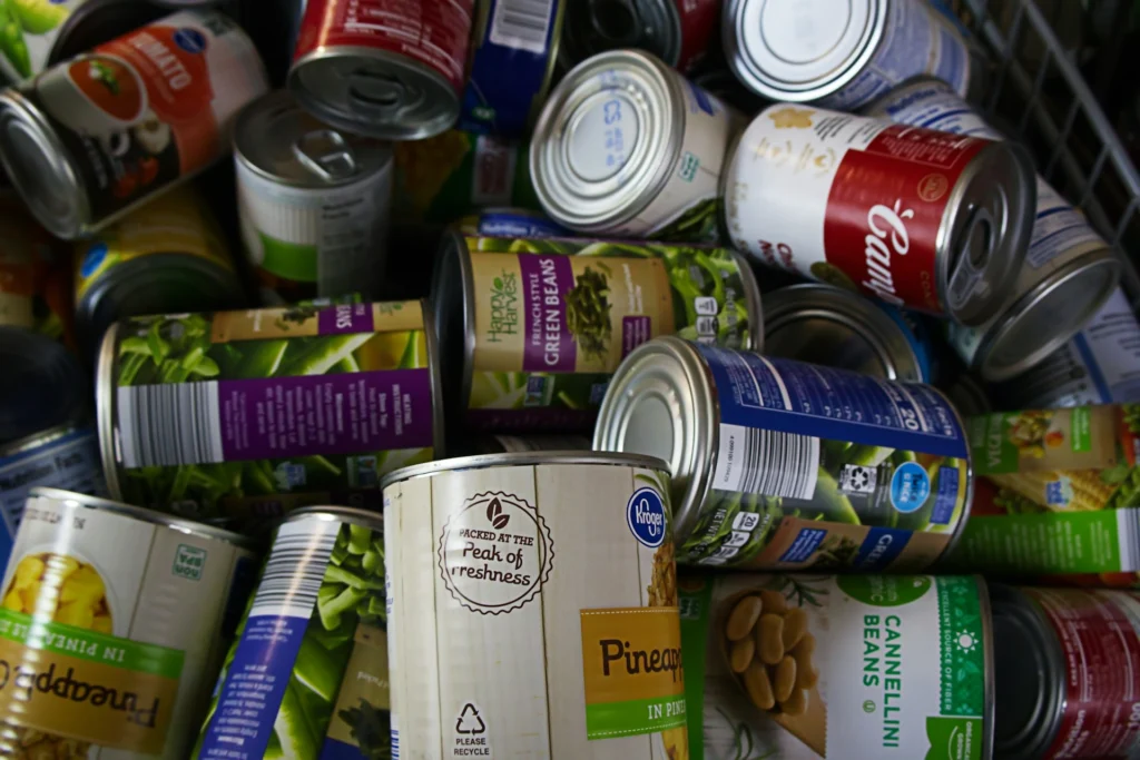 A pile of various canned goods, including beans, pineapple, and soup, with colorful labels, creating a cluttered but vibrant and resourceful scene.