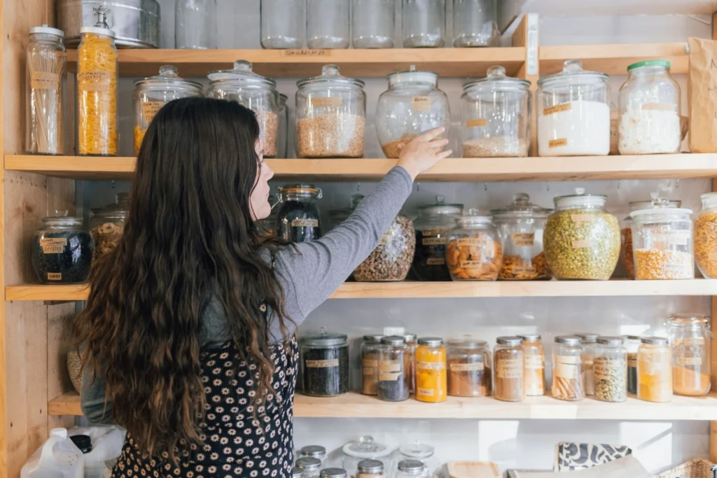 A woman with long brown hair reaches for a jar on a wooden shelf filled with various labeled jars of grains and spices, creating an organized, homey feel.
