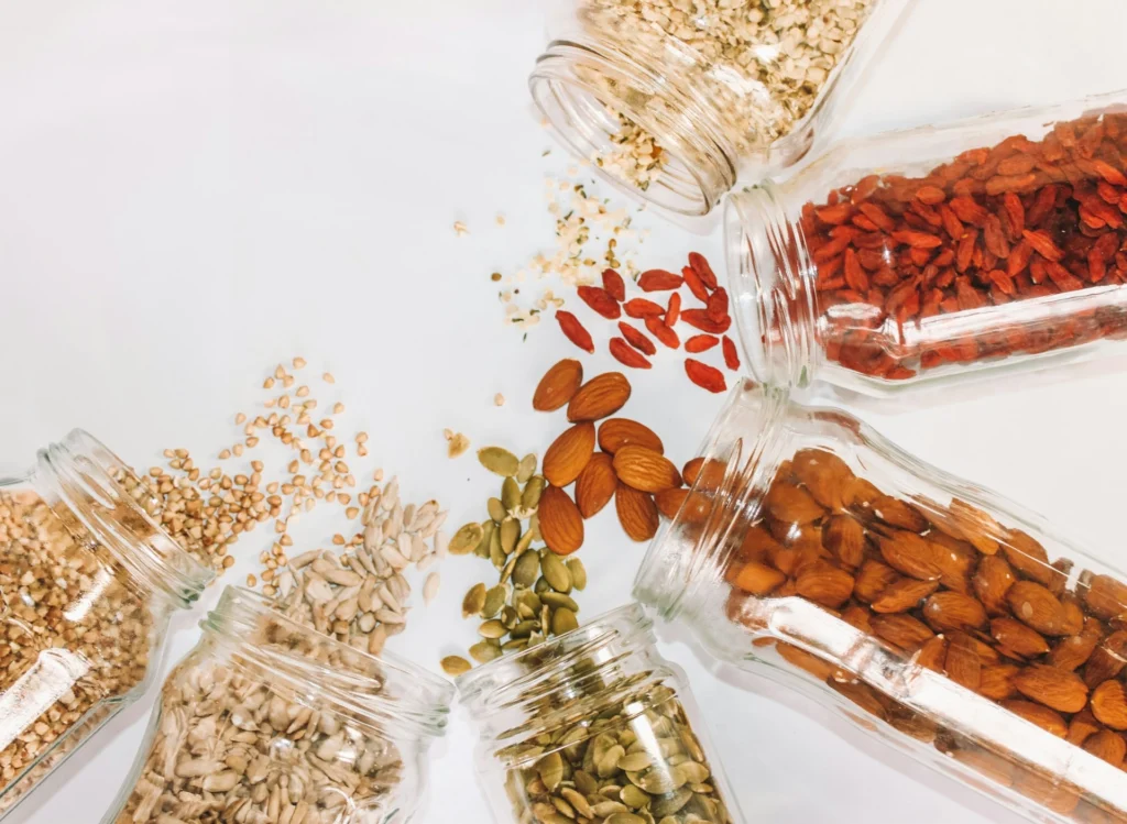 Various seeds and nuts, including almonds and goji berries, spill from glass jars arranged in a circle on a white surface, creating a vibrant, healthy display.