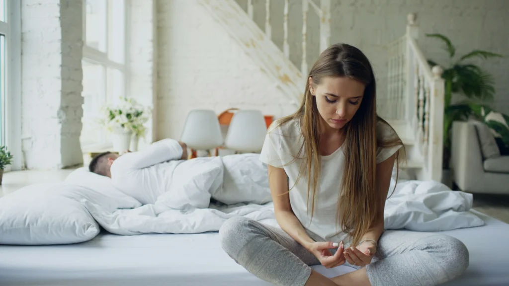 A woman looking unhappy while her partner is asleep, facing the other way, showing a sign that their relationship might need a cool-off