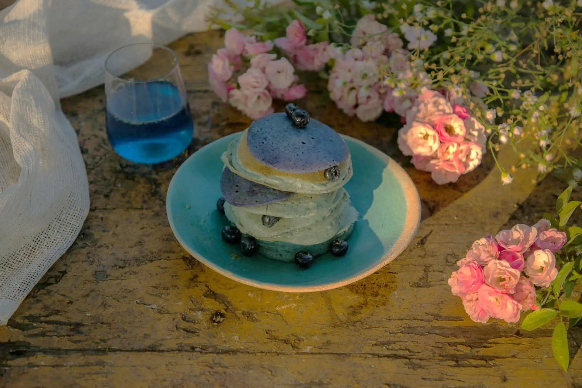 Stack of blue butterfly pea pancakes on a blue plate with a blue elixir drink, surrounded by roses and flowers on a wooden table