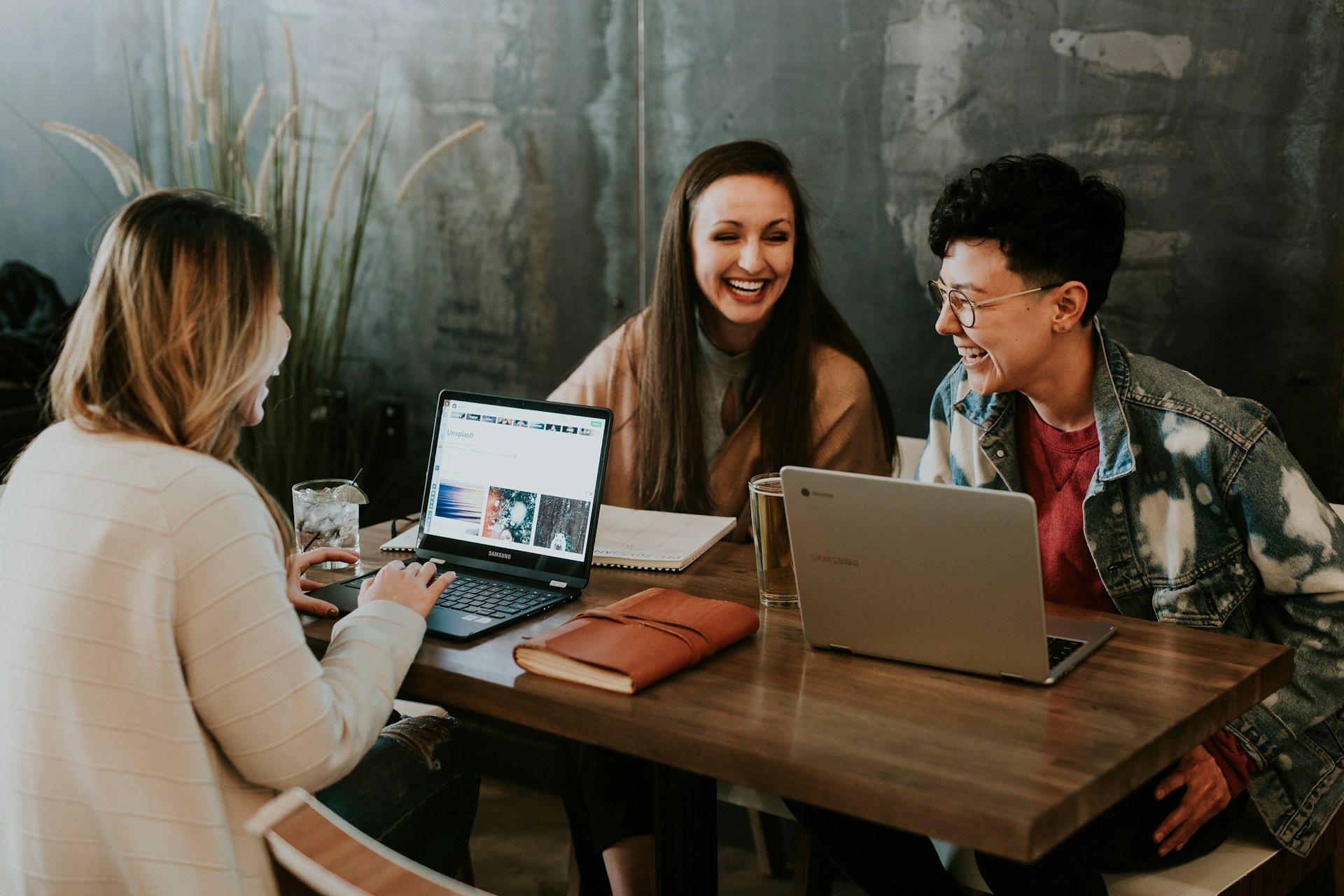 Three people are sitting at a wooden table with laptops, laughing and engaged in conversation, creating a warm and collaborative atmosphere.