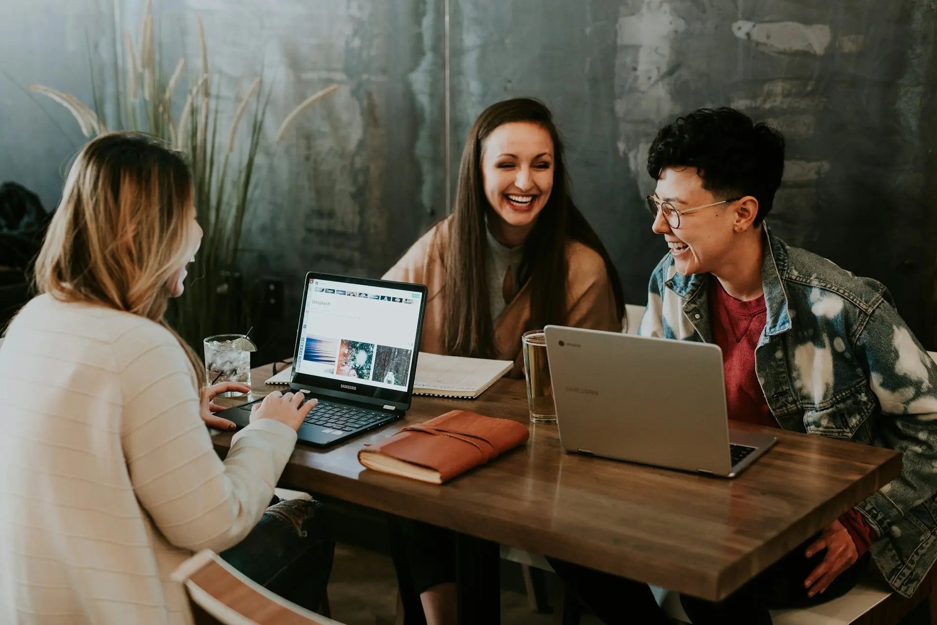 Three people are sitting at a wooden table with laptops, laughing and engaged in conversation, creating a warm and collaborative atmosphere.