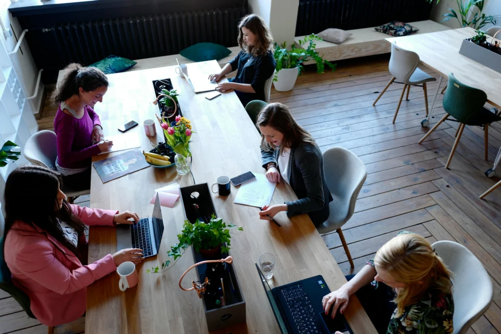 Aerial view of five women working at a long wooden table in a bright, modern office. They use laptops and take notes, surrounded by potted plants.