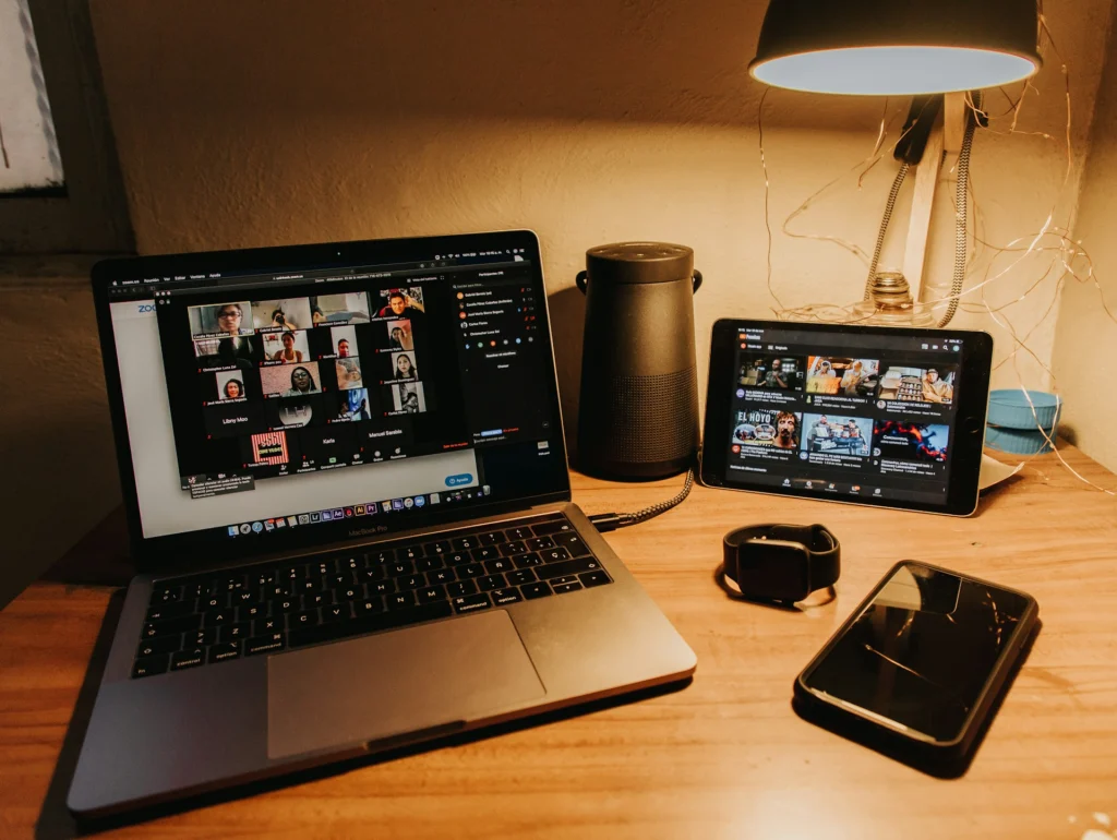 A cozy workspace with a laptop displaying a video call, a tablet showing video thumbnails, a smart speaker, a smartwatch, and a smartphone on a wooden desk.