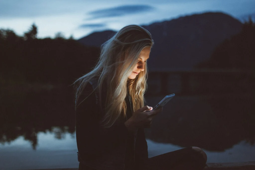 A woman seated on a dock, focused on her phone, with water and boats visible in the background.