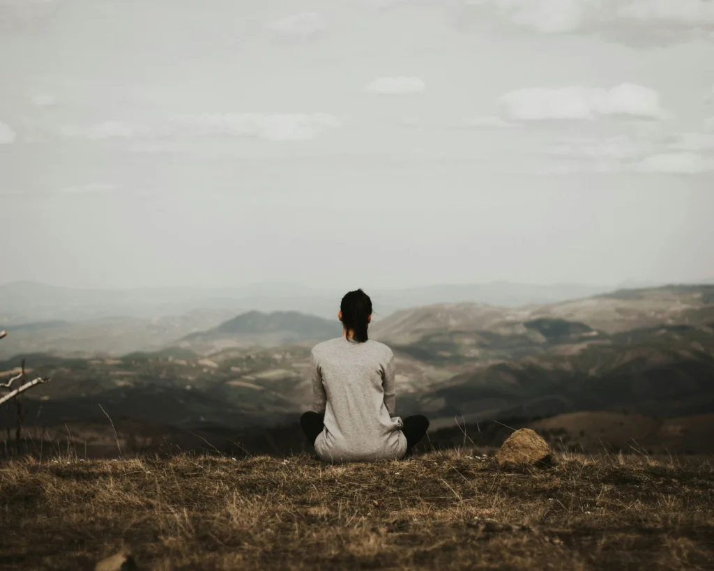 A woman relaxes on a hilltop, overlooking a picturesque valley filled with trees and rolling hills under a clear sky.