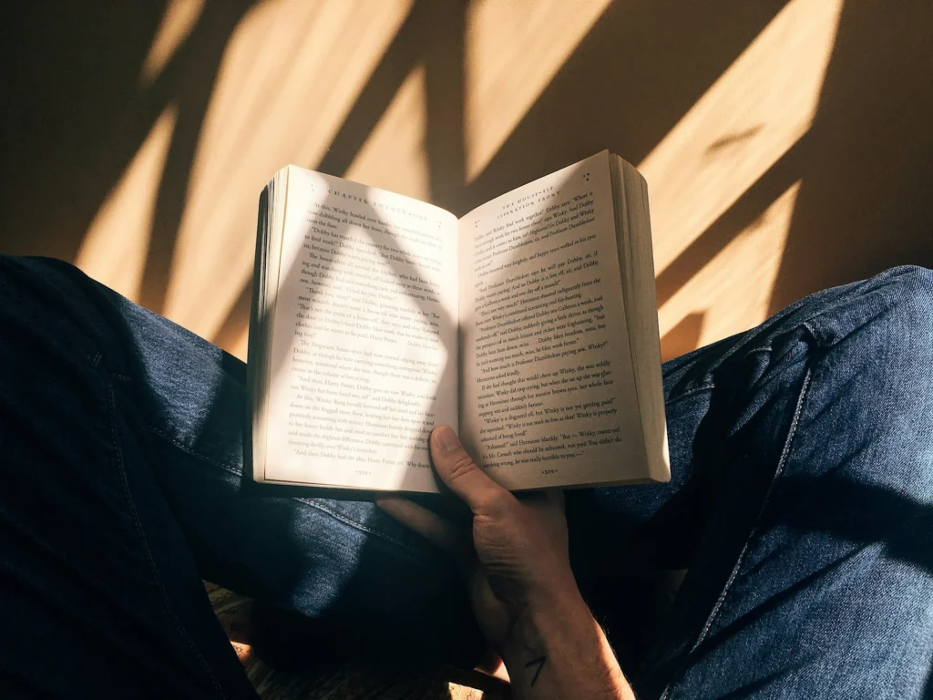 Individual wearing blue jeans cross-legged on the floor, engrossed in an open book.