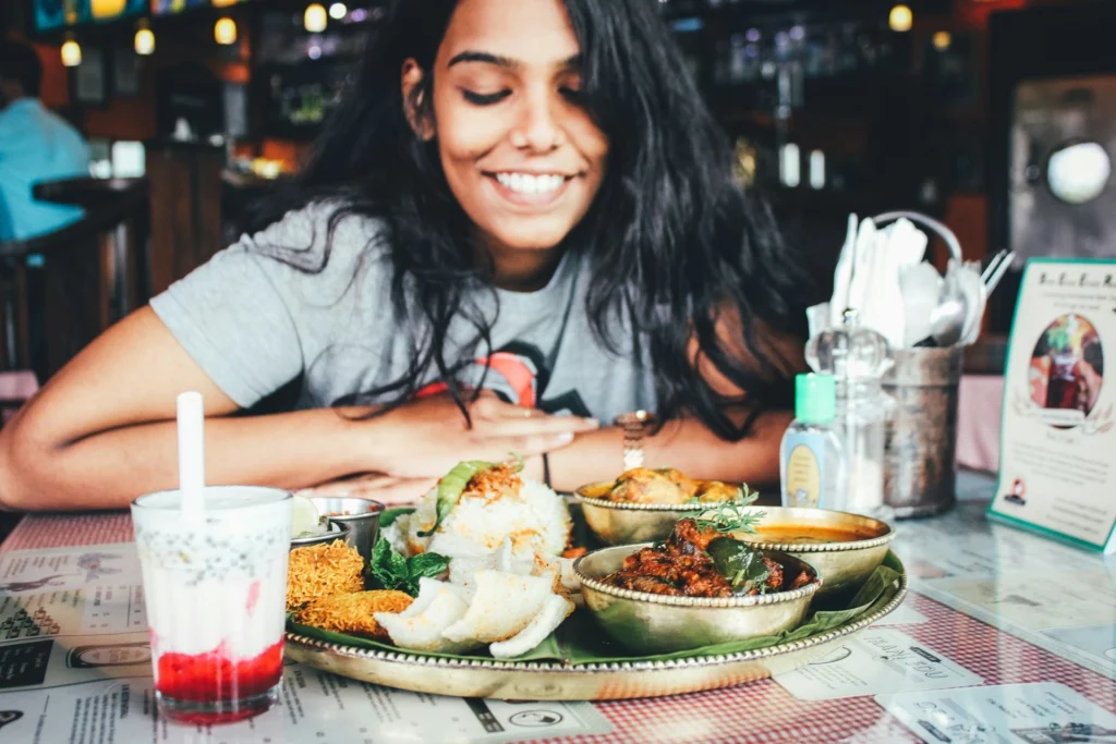 Smiling woman in a gray shirt admires a platter of diverse Indian dishes and a pink drink with a straw, conveying joy and anticipation.