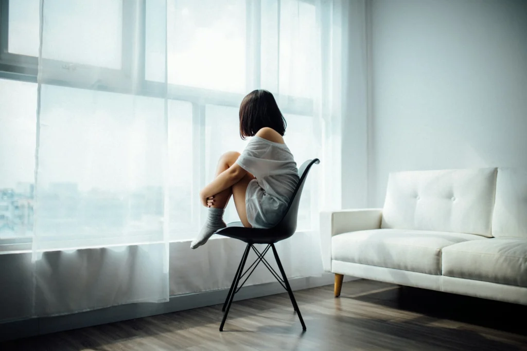 Person sitting on a chair, gazing out a large window with sheer curtains. Light floods the room with a sofa to the side, creating a serene, contemplative mood.
