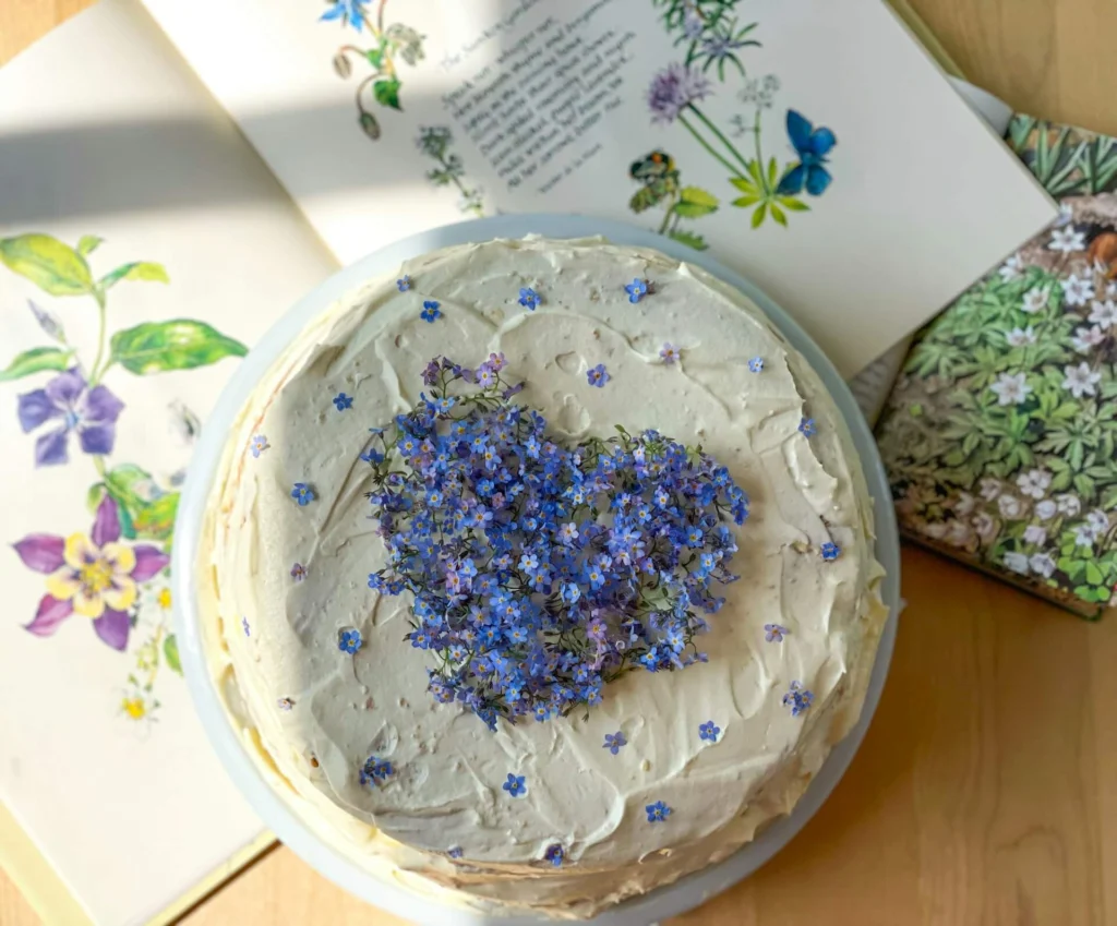 A round cake with cream icing decorated with purple flowers arranged in a heart shape