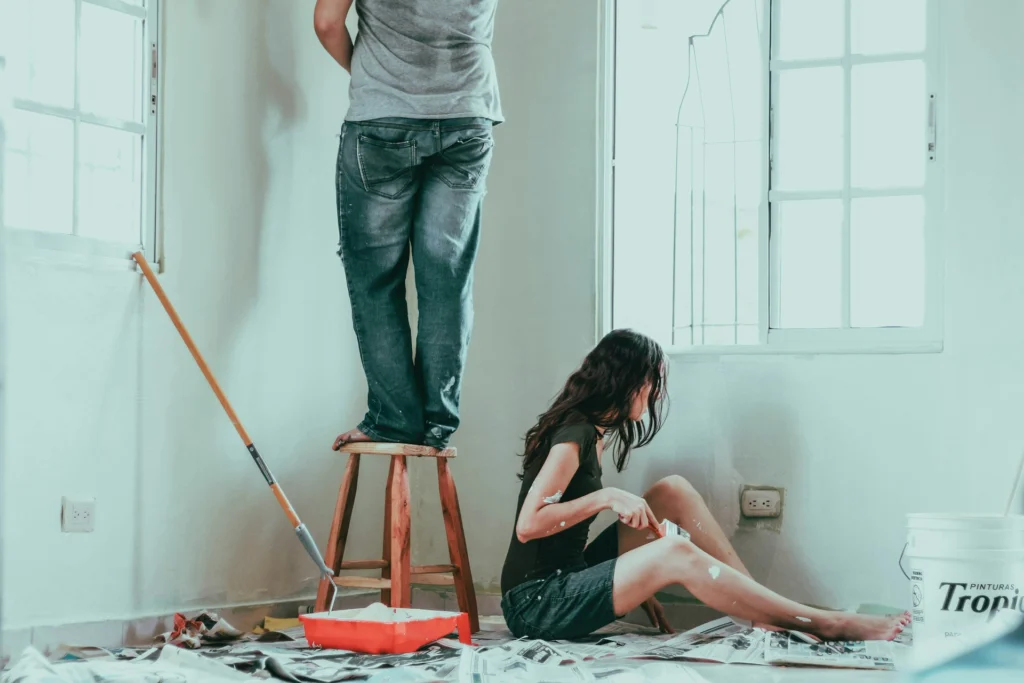 Two people paint a bright room. One stands on a stool while the other, seated on the floor, dips a brush into a paint bucket. A relaxed, collaborative mood.