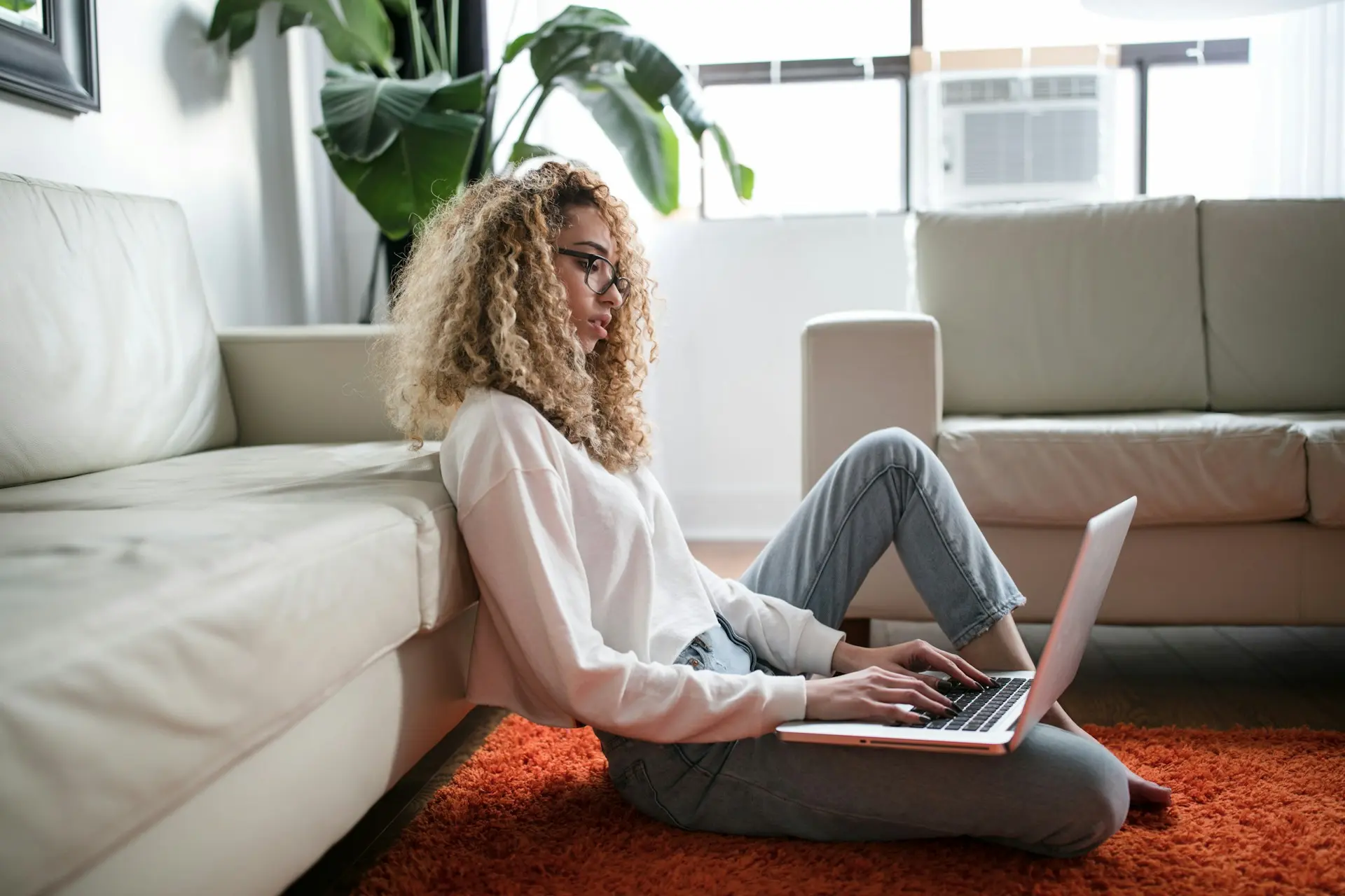 A woman with curly hair and glasses sits on an orange rug, working on a laptop. She's relaxed, leaning against a cream sofa in a bright room with plants.