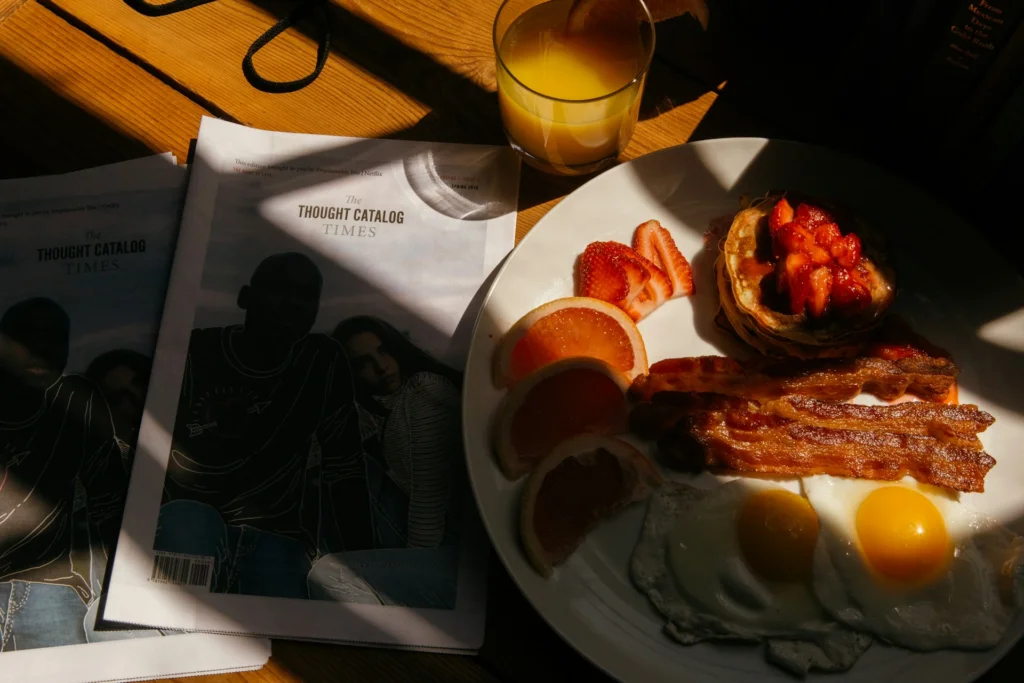 Breakfast scene with sunny-side-up eggs, bacon, grapefruit slices, strawberries, and waffles on a plate. Nearby, a glass of orange juice and two magazines titled "Thought Catalog Times" rest on a sunlit wooden table.