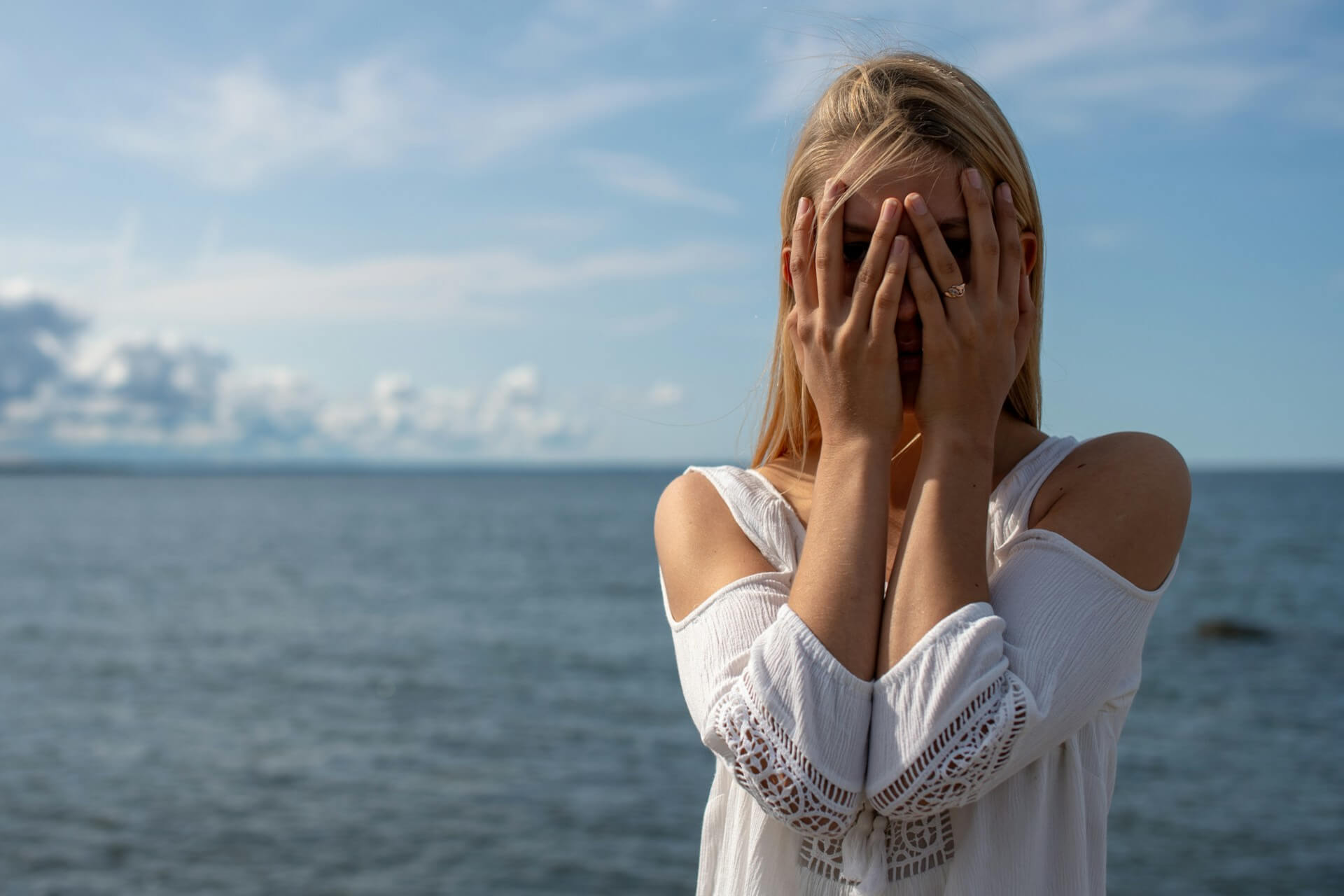 Woman in a white summer dress standing on seaside rocks by the ocean, hands raised to her face