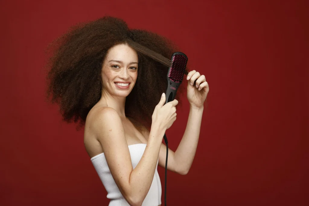 A woman with voluminous, tightly coiled curly hair uses a heated styling brush on a strand, straightening it out.