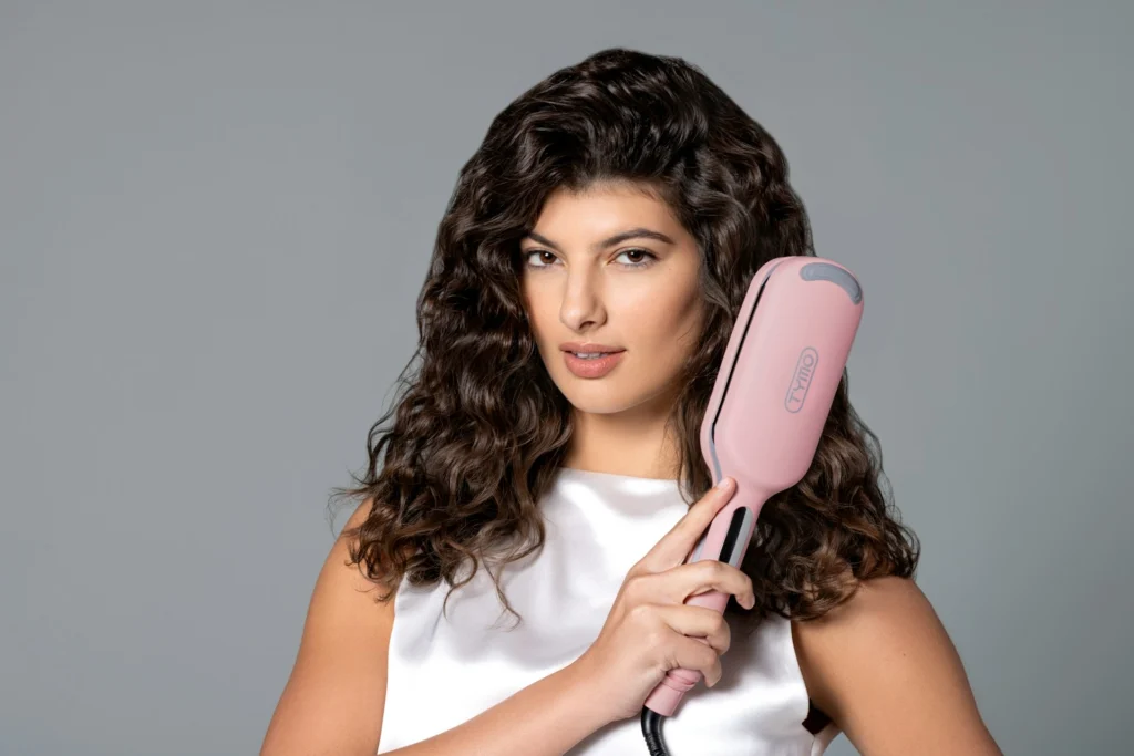 A woman with medium-length, wavy, brown hair holds a pink hair thermal brush and wears a shiny white top.