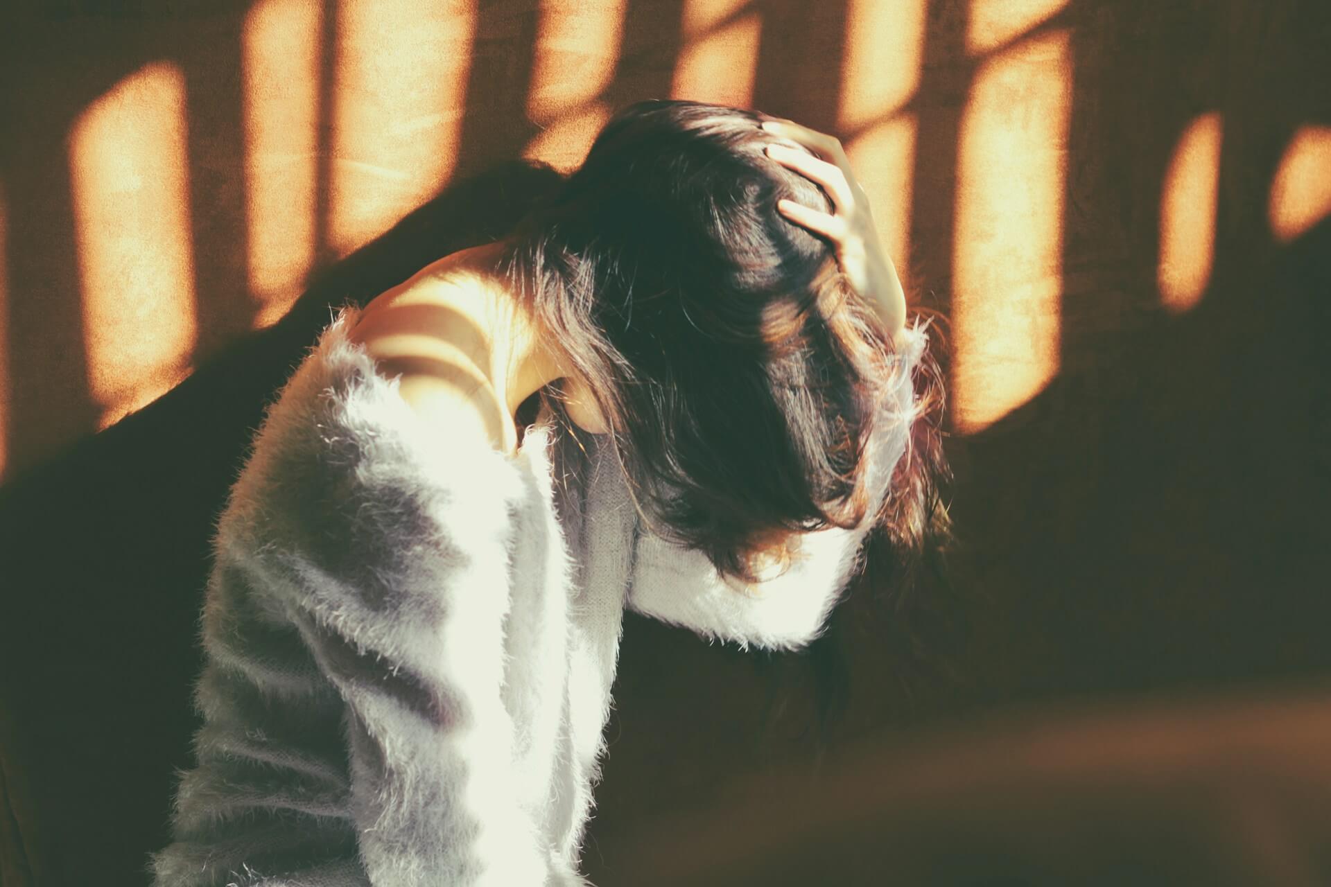 Woman in a white fur jumper leans against a sunlit wall, holding her head with one hand
