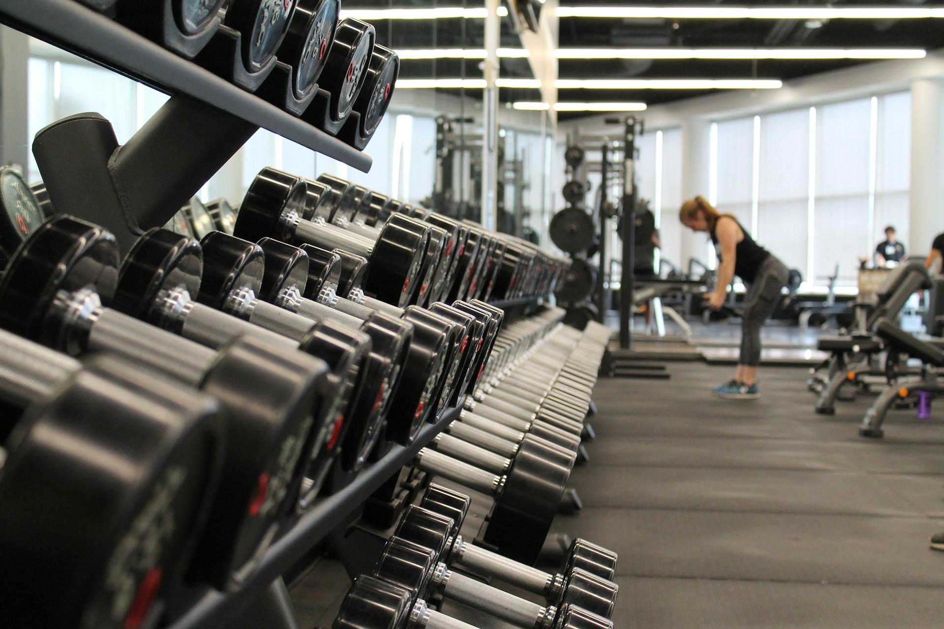 A gym interior featuring a row of dumbbells with a woman exercising in the background.