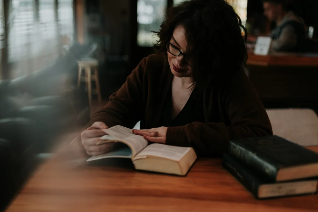 A woman sits at a table, engrossed in reading a book, with a focused expression on her face.