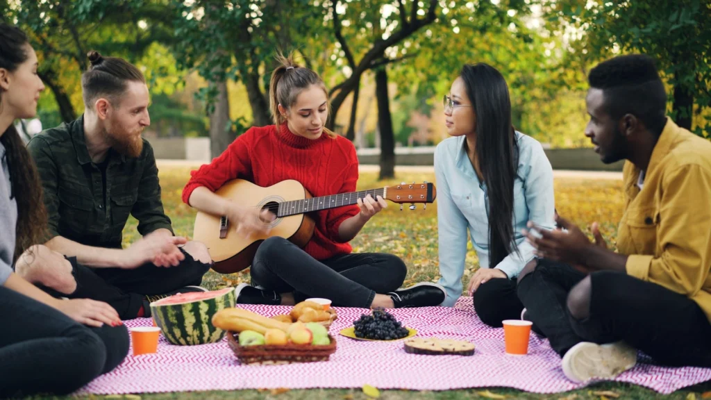 A group of young people sitting on a blanket, enjoying music while playing guitar together outdoors.