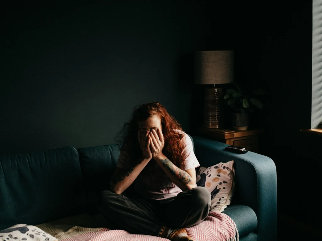 Red-haired woman sits cross-legged on a dark sofa, covering her face with both hands in a dim, quiet room, conveying distress or sadness.