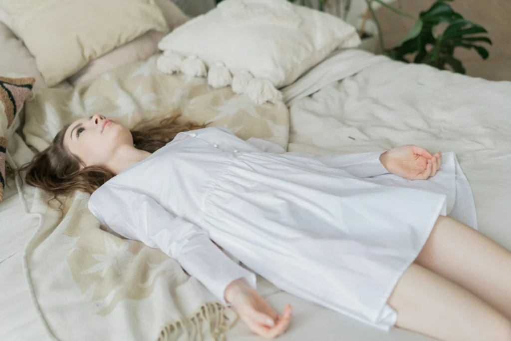 Young woman in a white dress lies on a cozy bed among pillows, gazing upward with a calm, dreamy expression in soft natural light.