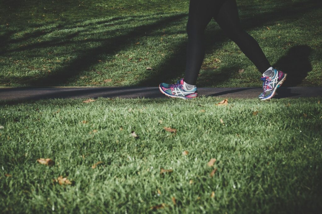 Person in black leggings walks along a paved park path wearing colorful running shoes, casting a long shadow over sunlit grass and fallen leaves.