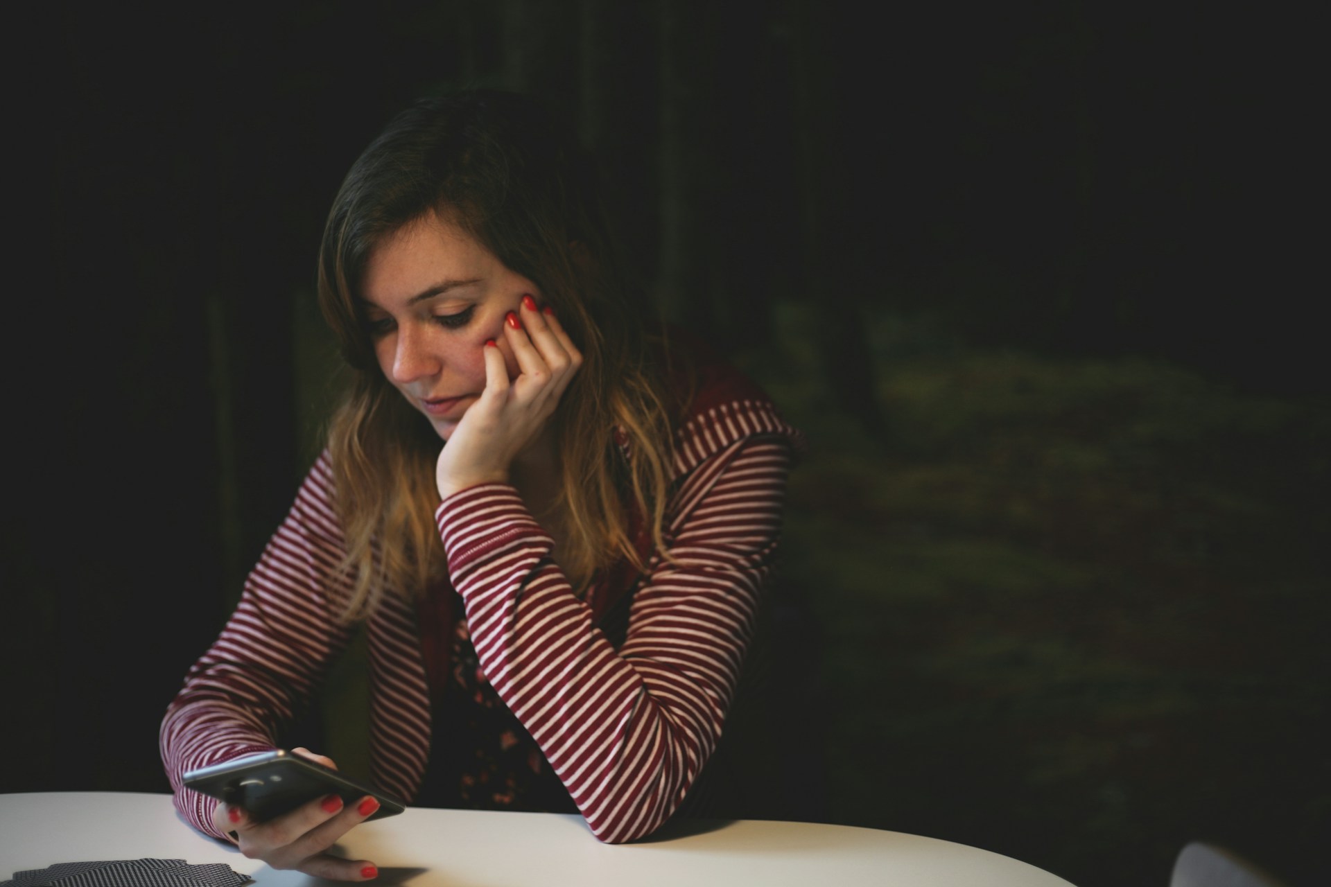 A woman sitting at a table, looking at her cell phone, with soft lighting and minimal decor in the background.