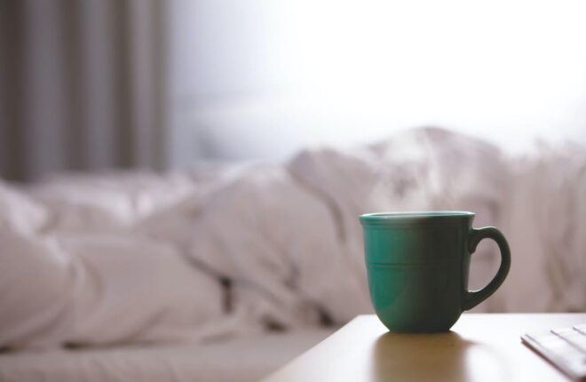hot mug of coffee on end table by empty bed one of the benefits of waking up at 5 am