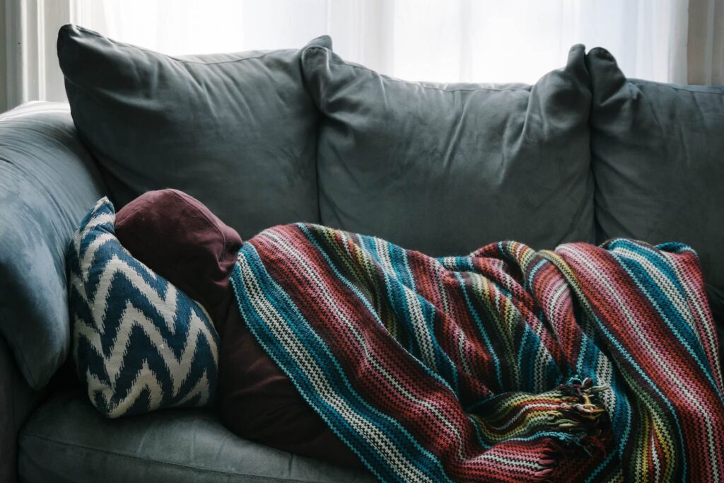 A person lays on a gray couch, facing the back cushions. They wear a red hoodie with the hood pulled up over their head. They're laying on a blue and white chevron striped pillow and underneath a multi-color woven blanket with tassles - how to recover from the flu