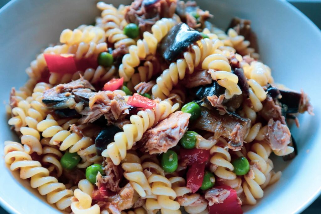A white bowl holds a mixture of spiral pasta, chopped tomatoes, peas and some kind of seafood.