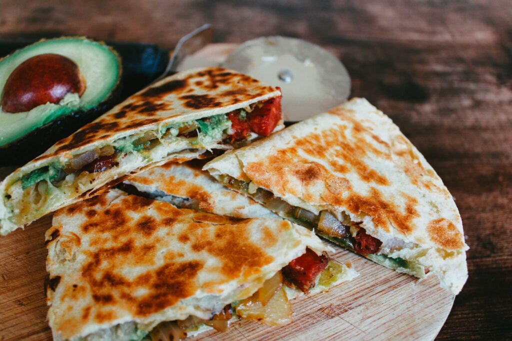 A round wooden board sits on a dark wooden countertop. Two quesadillas sliced in half sit on the board along with half an avocado. A pizza cutter is in the background. The quesadillas contain various fried vegetables and cheese.