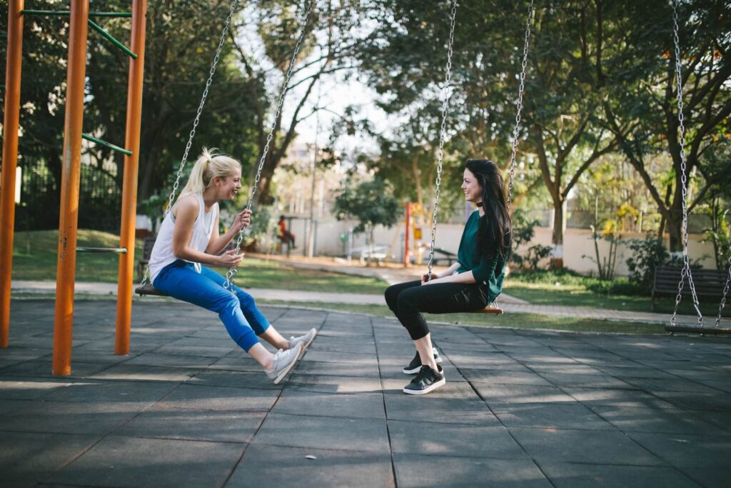 Two white women sit on swings at a layground. One has a blonde ponytail, a white tank top, blue jans and gray tennis shoes. The other has long brown hair, a longsleeve green shirt, black jeans and black tennis shoes. They're smiling at each other mid-conversation.
