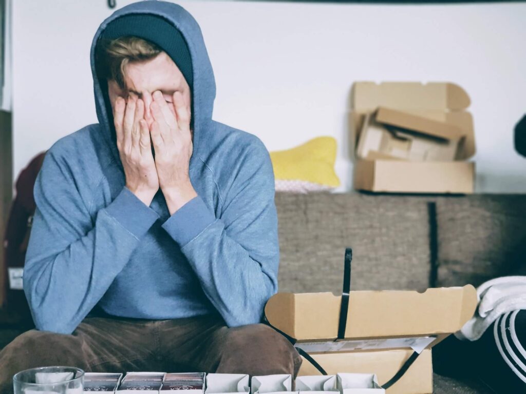 A white man sits on a gray couch and covers his face with both of his hands. He wears a blue hoodie and the hood is over his head. Empty boxes are all over the couch.